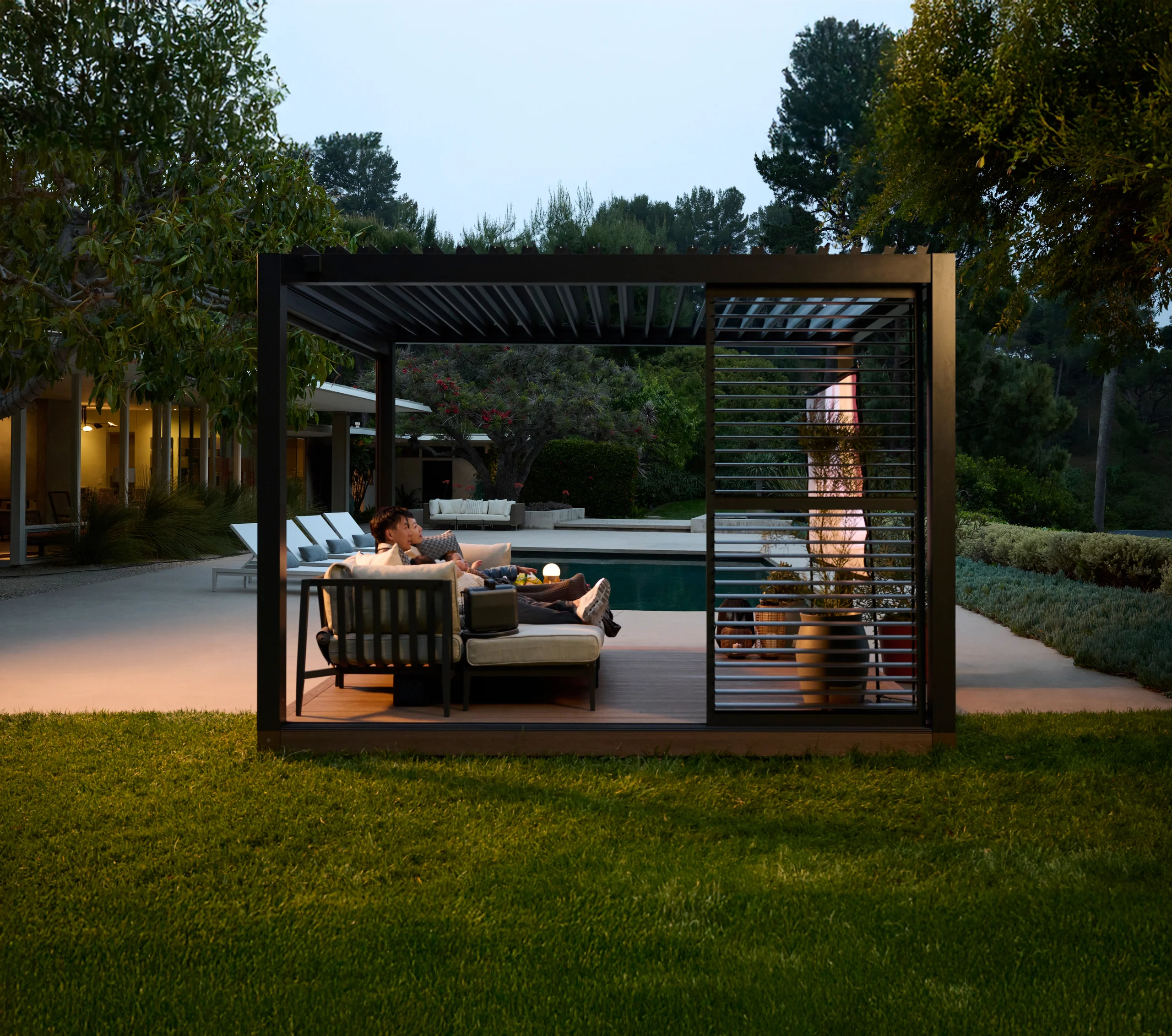 A couple relaxing in a Haven pavilion with louvered privacy panels beside a pool at dusk